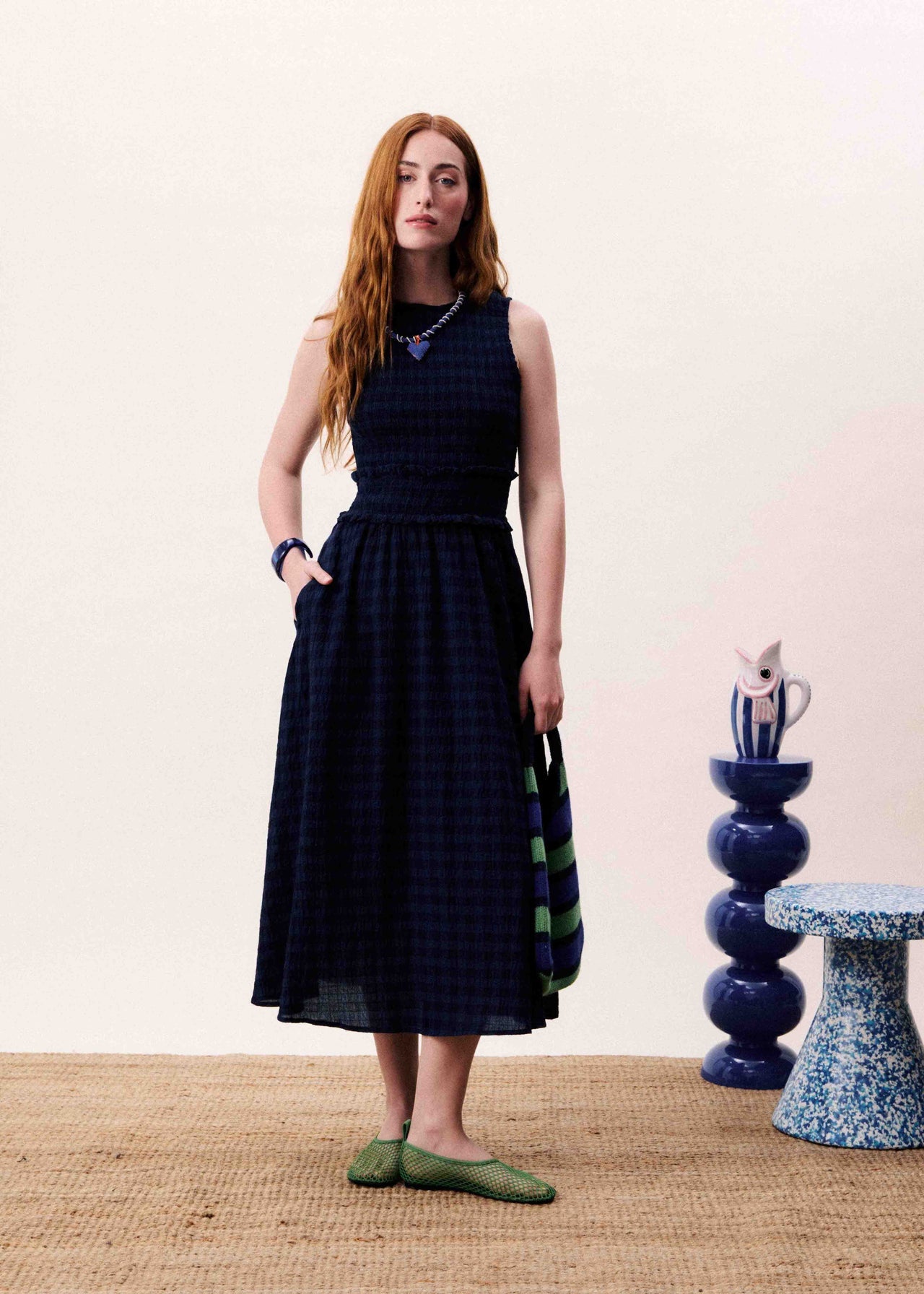 Woman wearing a dark blue dress with a pattern, standing in a room with a pink wall and decorative items.