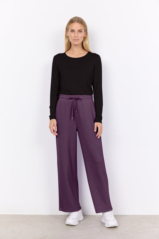 Woman wearing a black long-sleeve top and purple pants against a white background