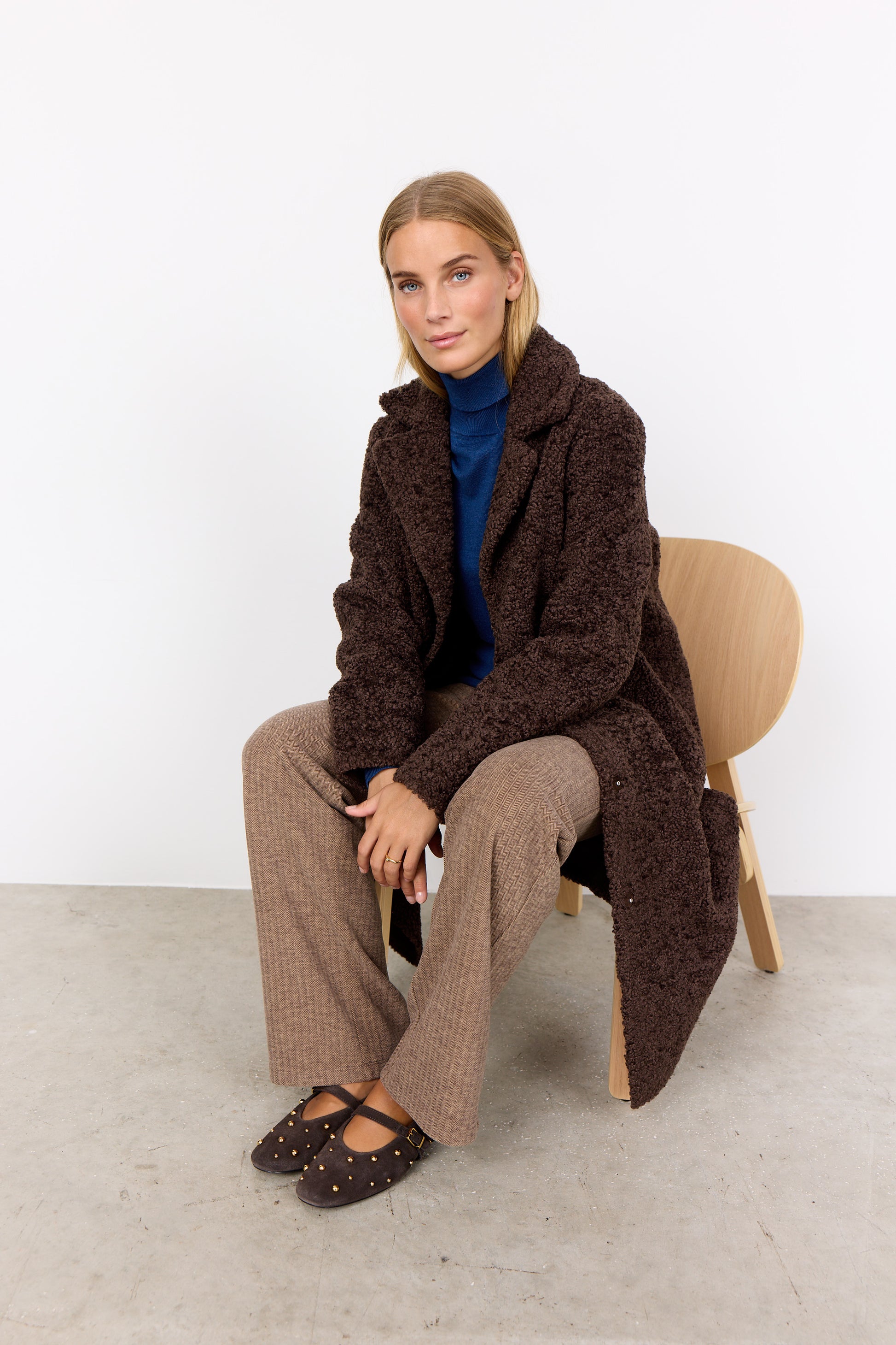 Woman sitting on a chair wearing a brown textured coat against a white background
