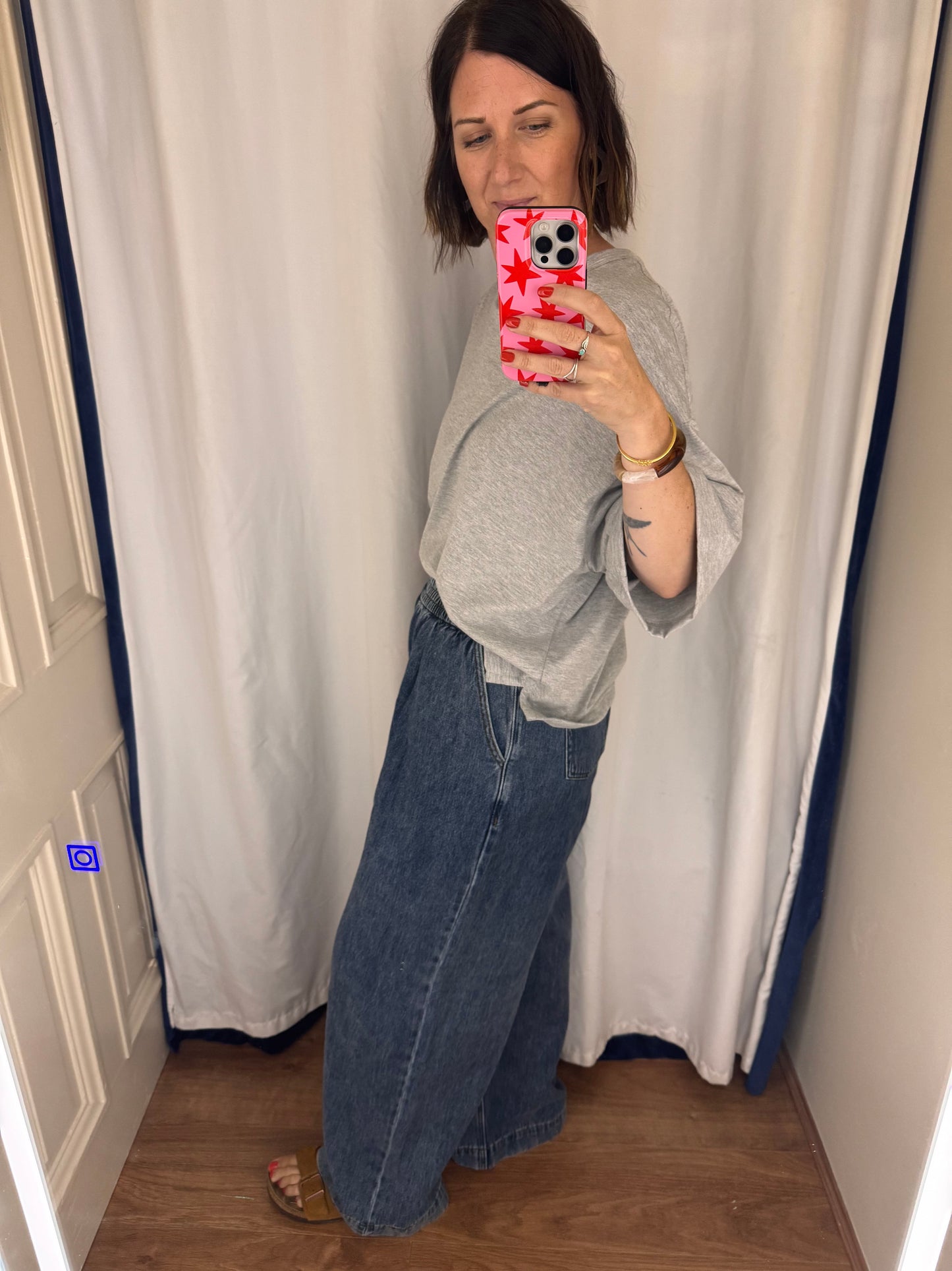 Person taking a mirror selfie in a changing room with a curtain and wooden furniture.