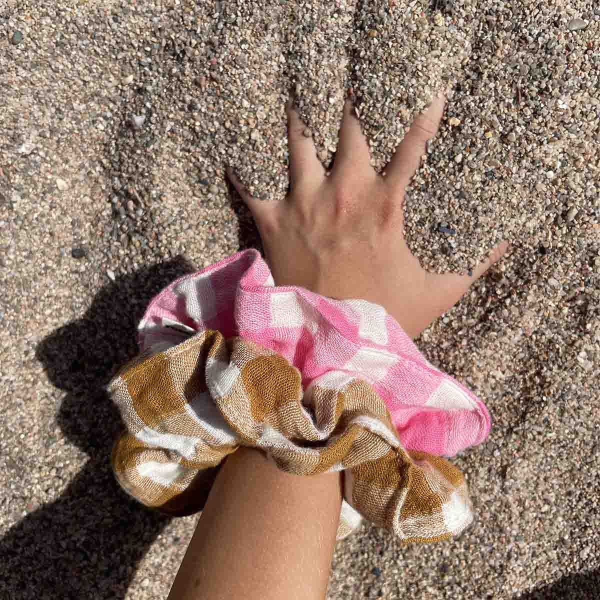 Hand reaching towards a pink and white checkered scrunchie on sandy ground