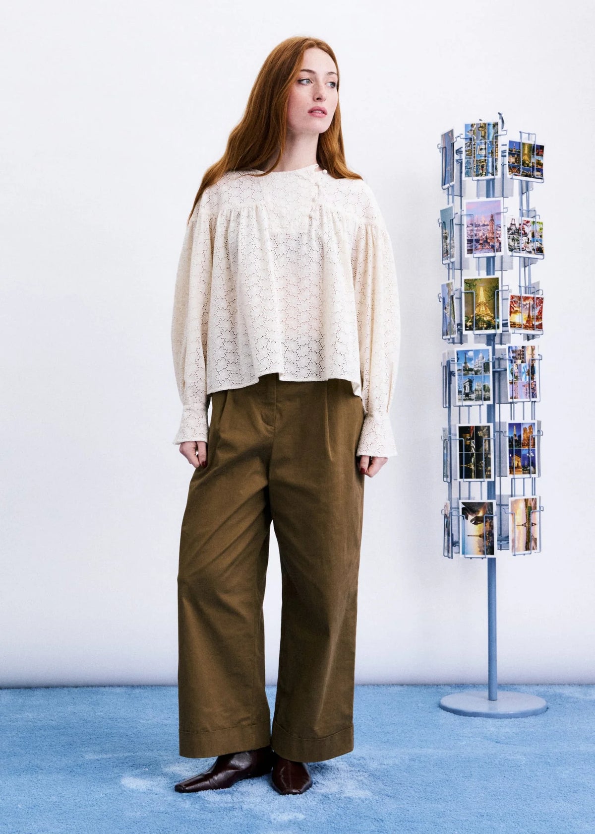 A woman stands in a studio wearing a cream lace-trim blouse and olive wide-leg trousers, with a blue floor and a tall rotating photo display stand beside her.
