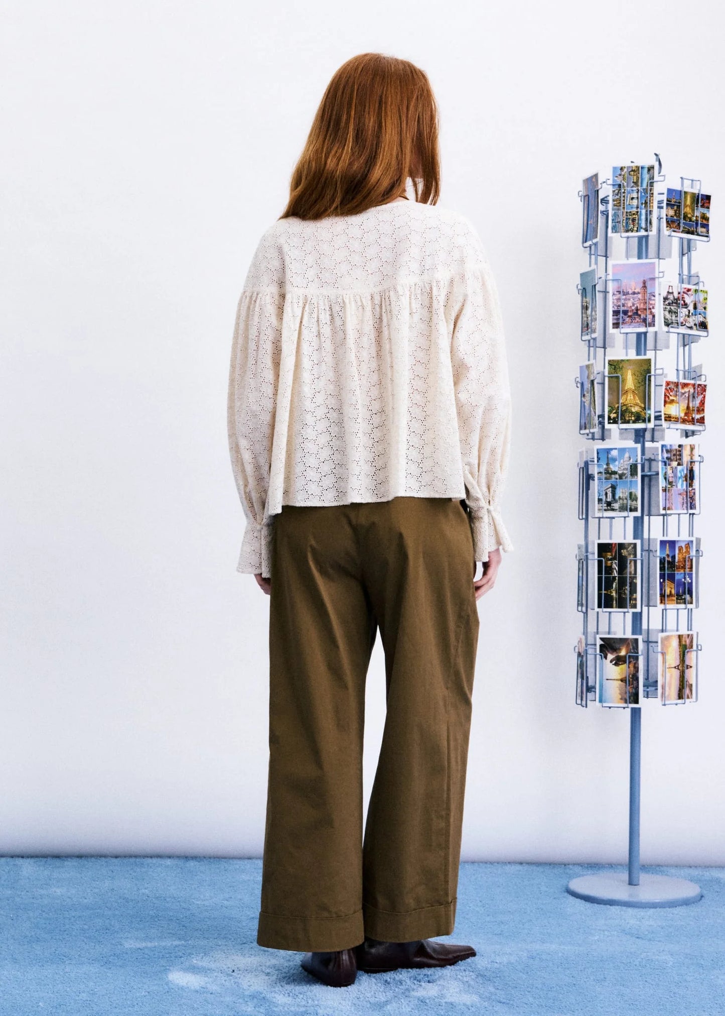 A woman stands with her back to the camera wearing a cream-colored textured blouse and wide-leg brown trousers, standing in front of a white wall with a blue floor. A clothing display rack is visible on the right.