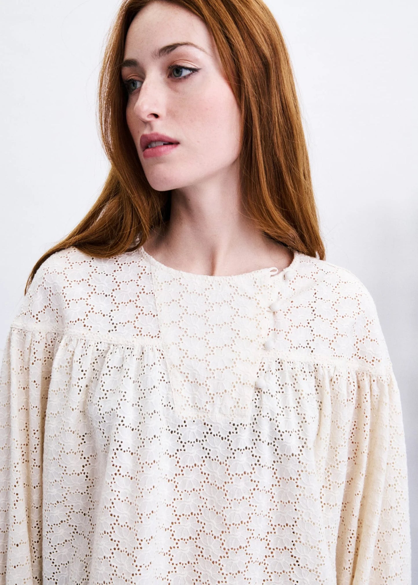 A close-up of a woman with red hair wearing a cream-colored eyelet lace top against a plain light background.