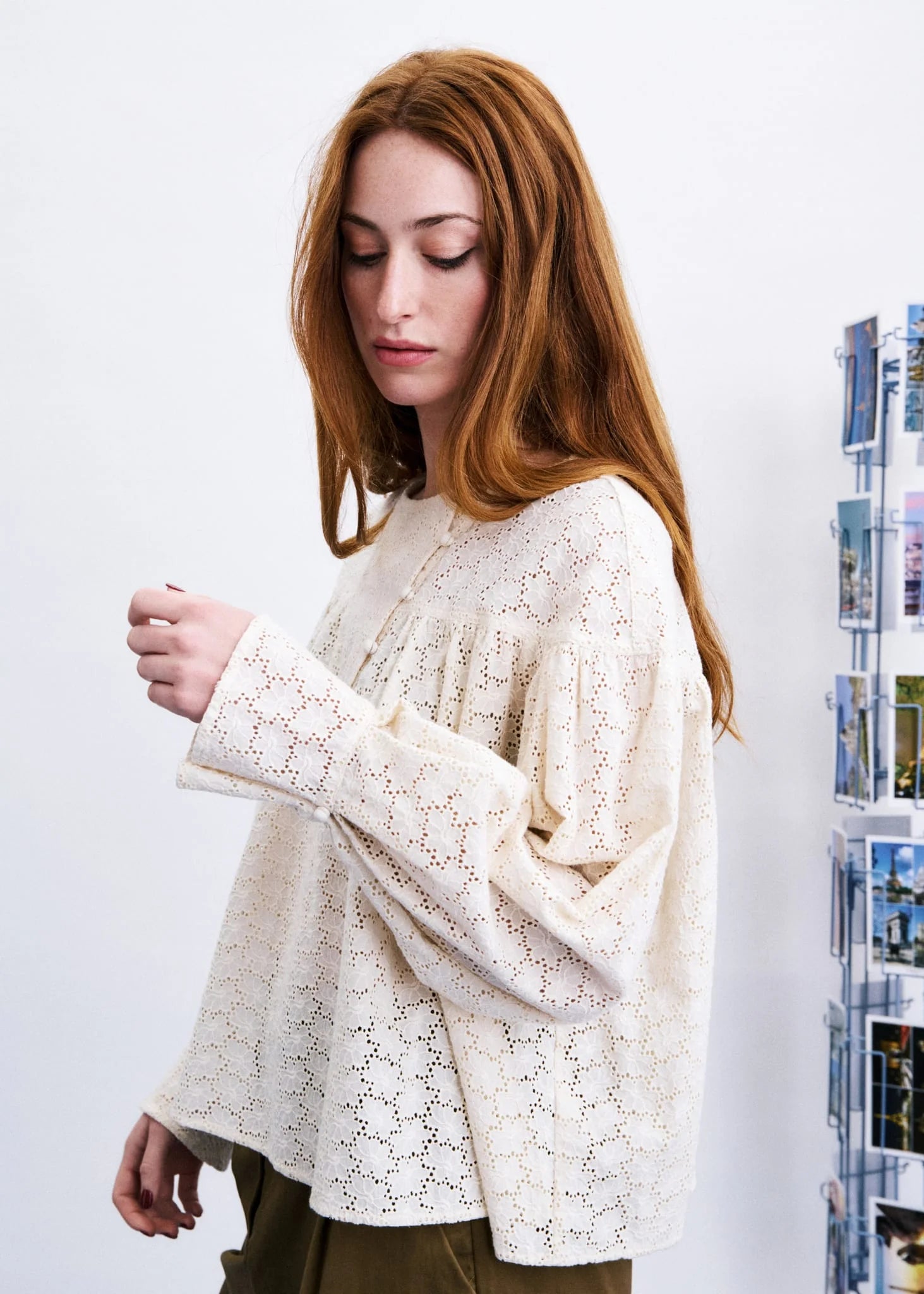 A woman with long red hair wearing a cream-colored, long-sleeved lace blouse, standing against a white wall next to a display rack.