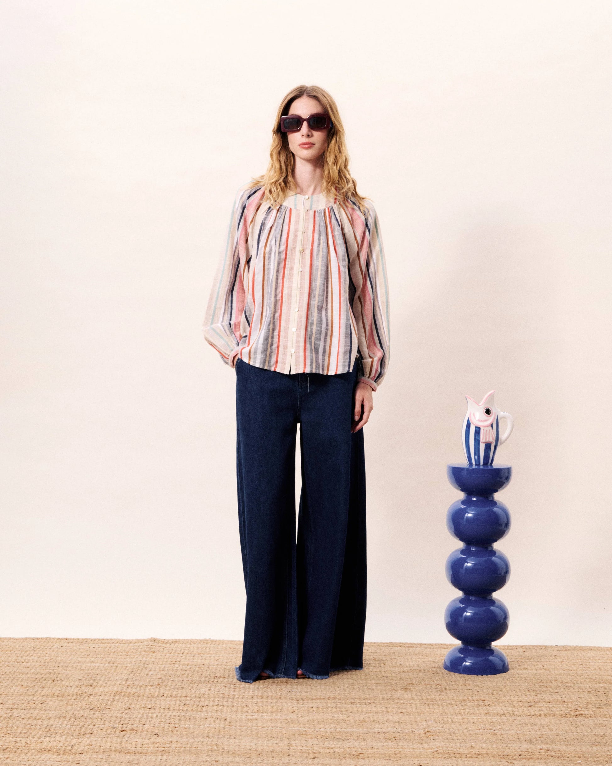 Woman wearing a colorful striped blouse and dark pants standing next to a blue decorative vase on a beige carpet.