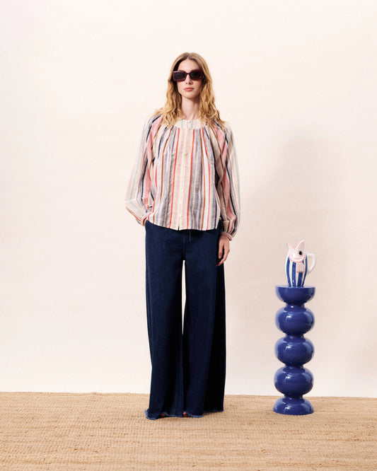 Woman wearing a colorful striped blouse and dark pants standing next to a blue decorative vase on a beige carpet.