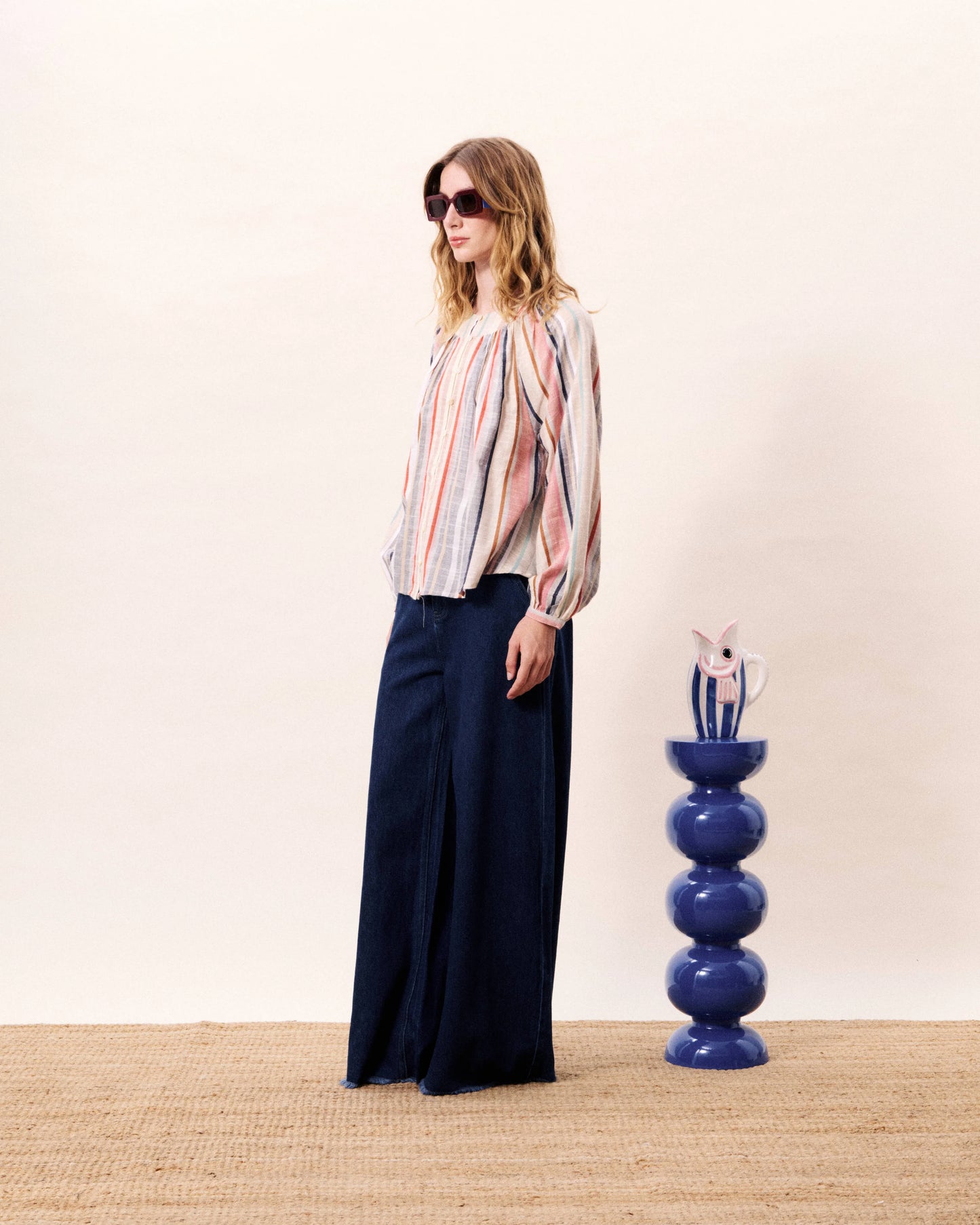 Woman wearing a colorful striped blouse and navy pants standing next to a blue decorative vase on a beige carpet.