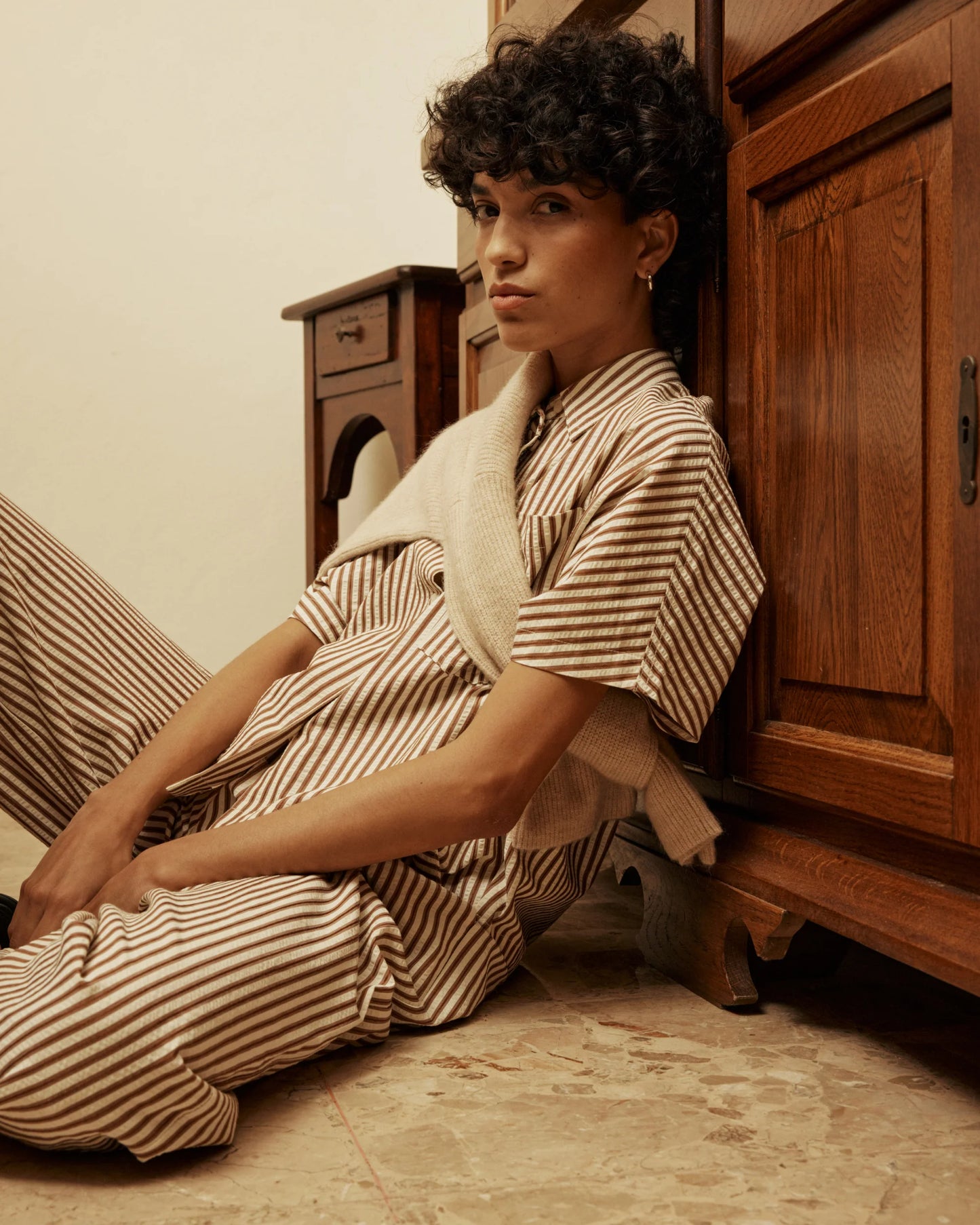 Person wearing a striped outfit sitting on the floor next to wooden furniture.