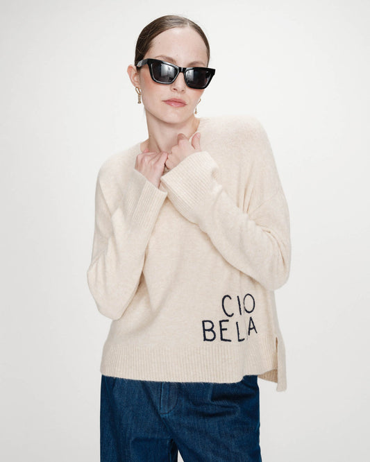 Person wearing a beige sweater with 'CIAO BELLA' text, dark sunglasses, and blue pants on a white background