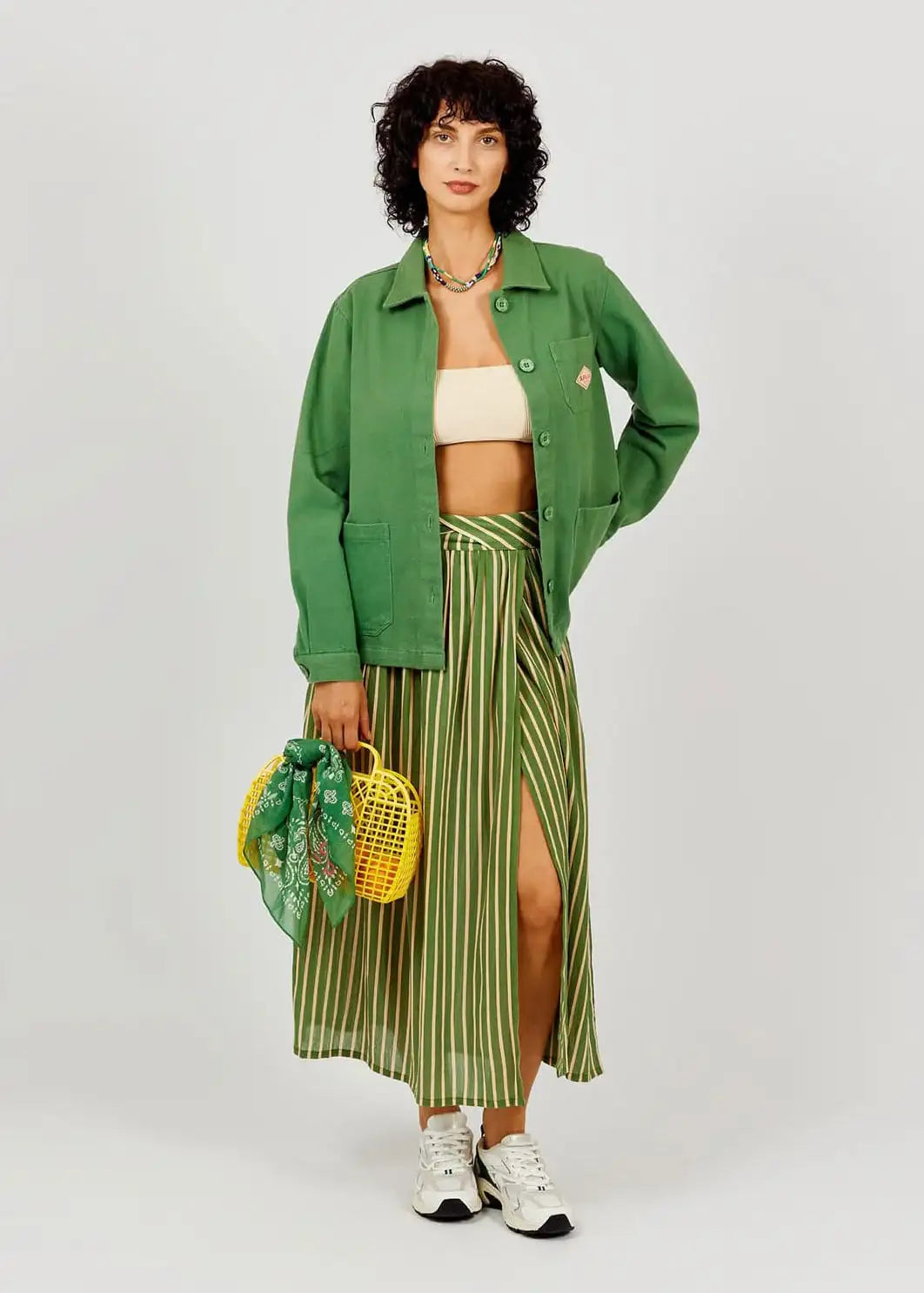 Woman wearing a green jacket and striped skirt holding a yellow bag on a white background