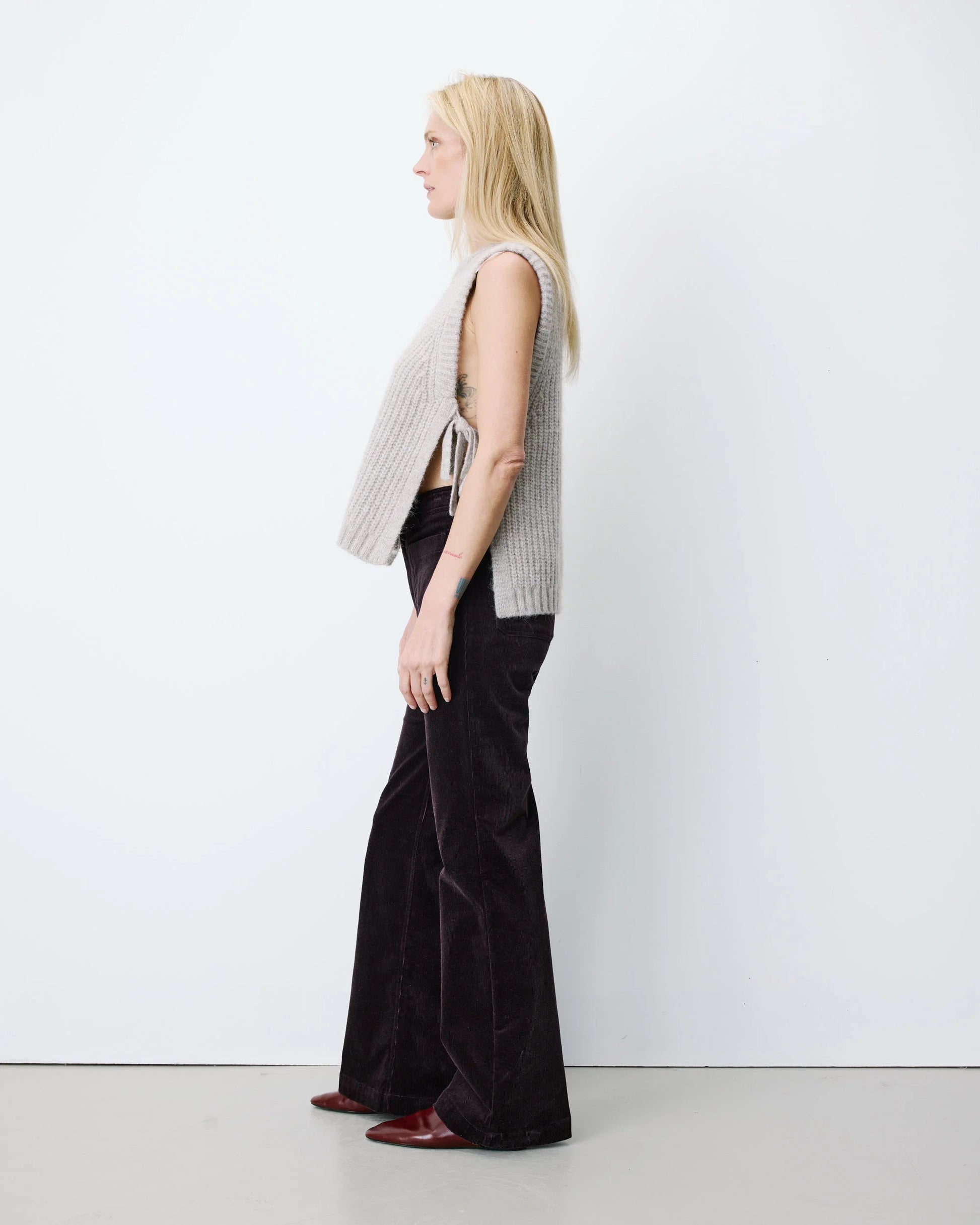 A blonde model shown in profile wearing a light gray ribbed sleeveless vest over a cropped black top and black wide-leg pants, standing against a plain white studio wall.