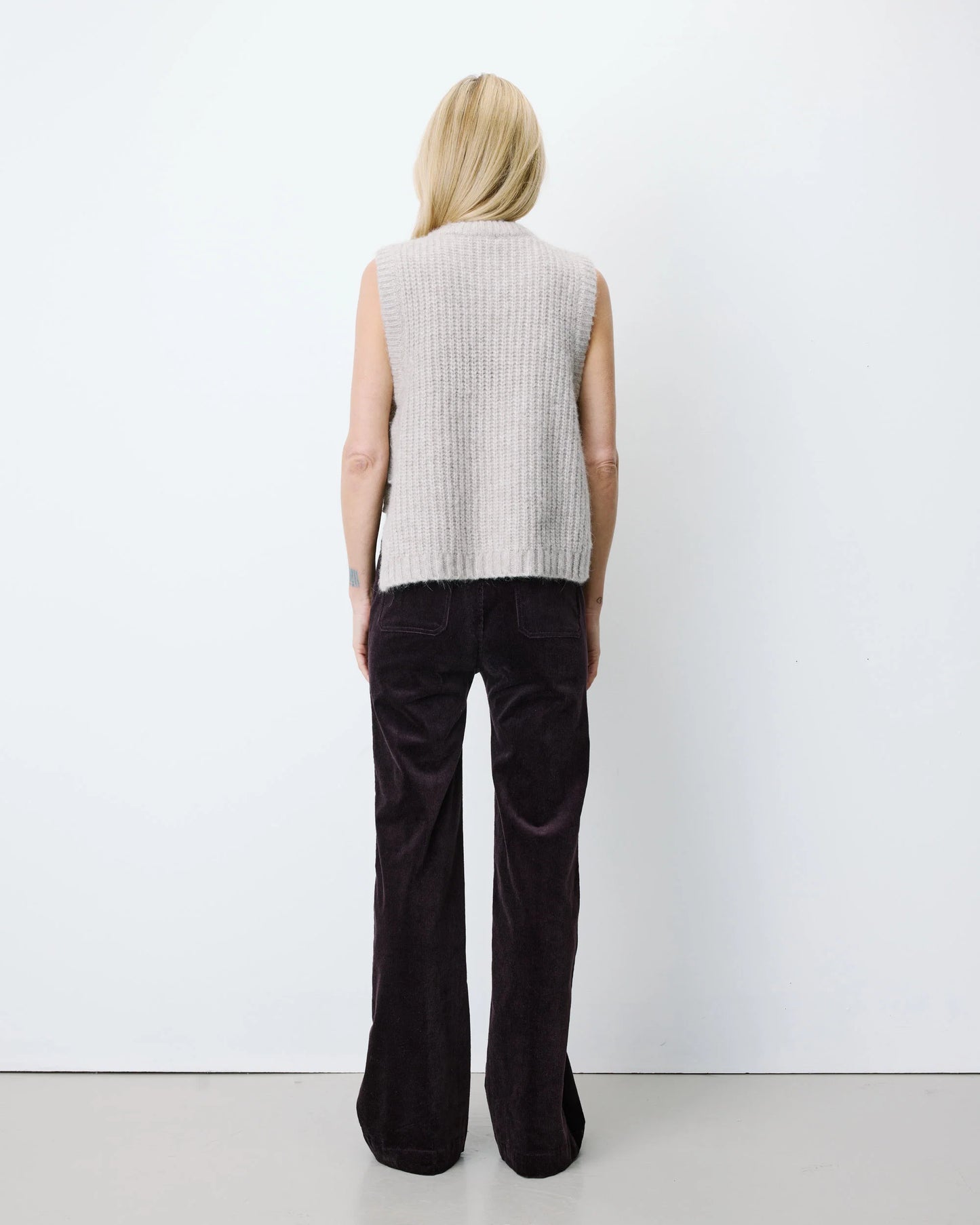 A woman stands with her back to the camera wearing a light gray ribbed knit sleeveless vest and dark brown wide-leg corduroy pants against a plain white studio background.