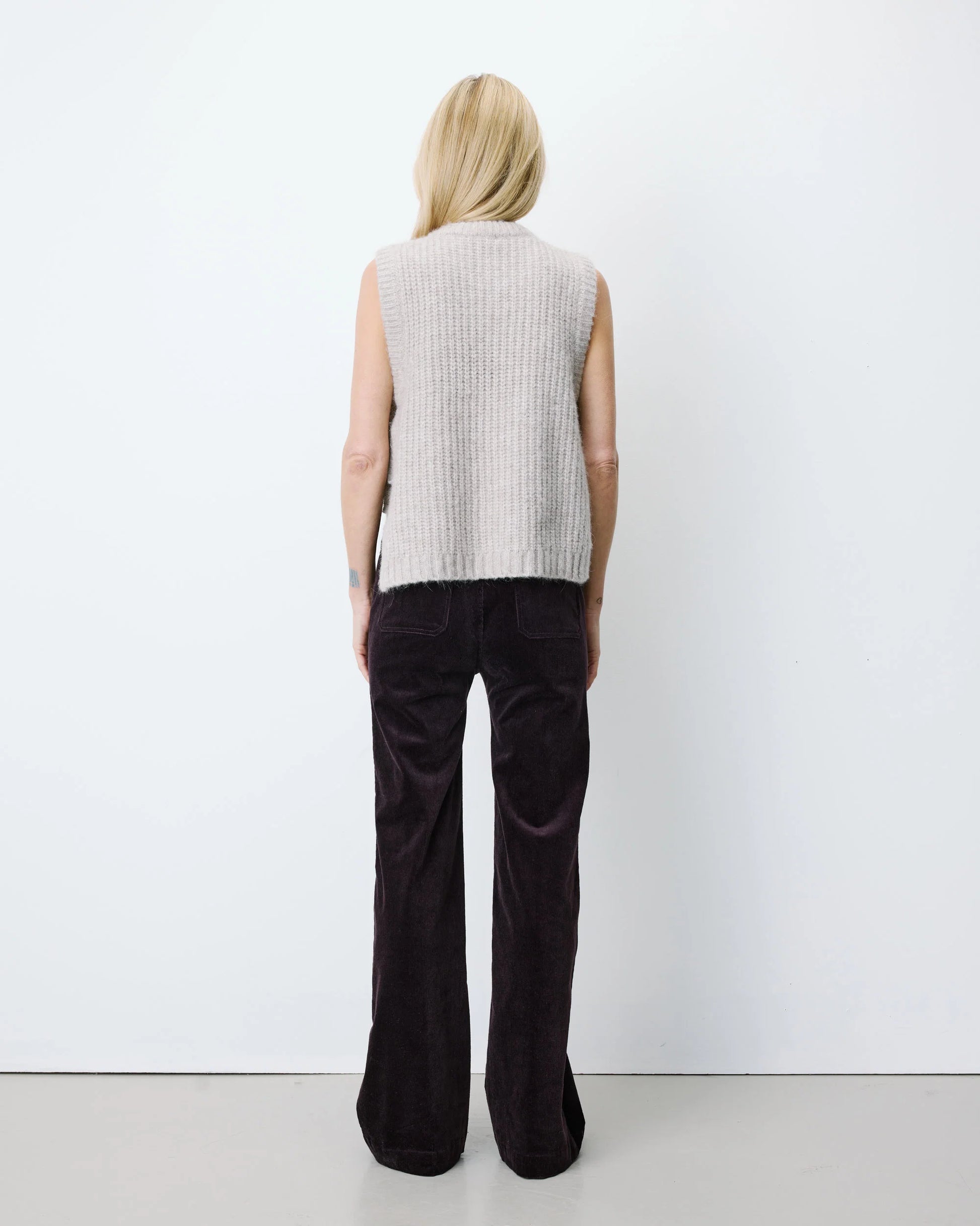 A woman stands with her back to the camera wearing a light gray ribbed knit sleeveless vest and dark brown wide-leg corduroy pants against a plain white studio background.
