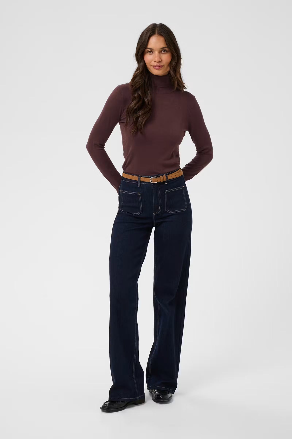 A woman modeling a dark brown mock-neck long-sleeve top and dark blue wide-leg jeans with a tan belt, standing against a light gray studio background.