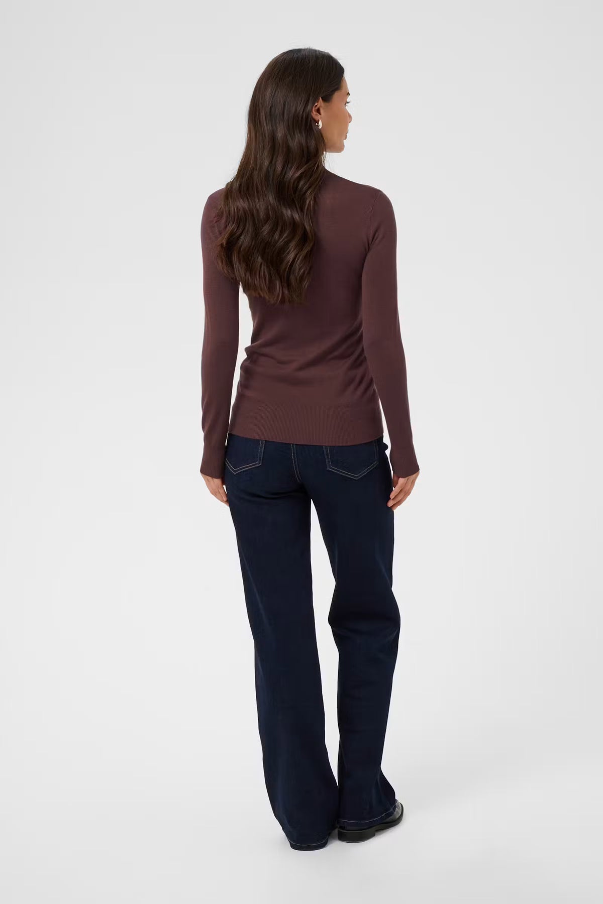 A woman shown from the back wearing a fitted long-sleeve ribbed top in a dark plum-brown color and dark navy blue jeans, standing against a plain light gray studio background.