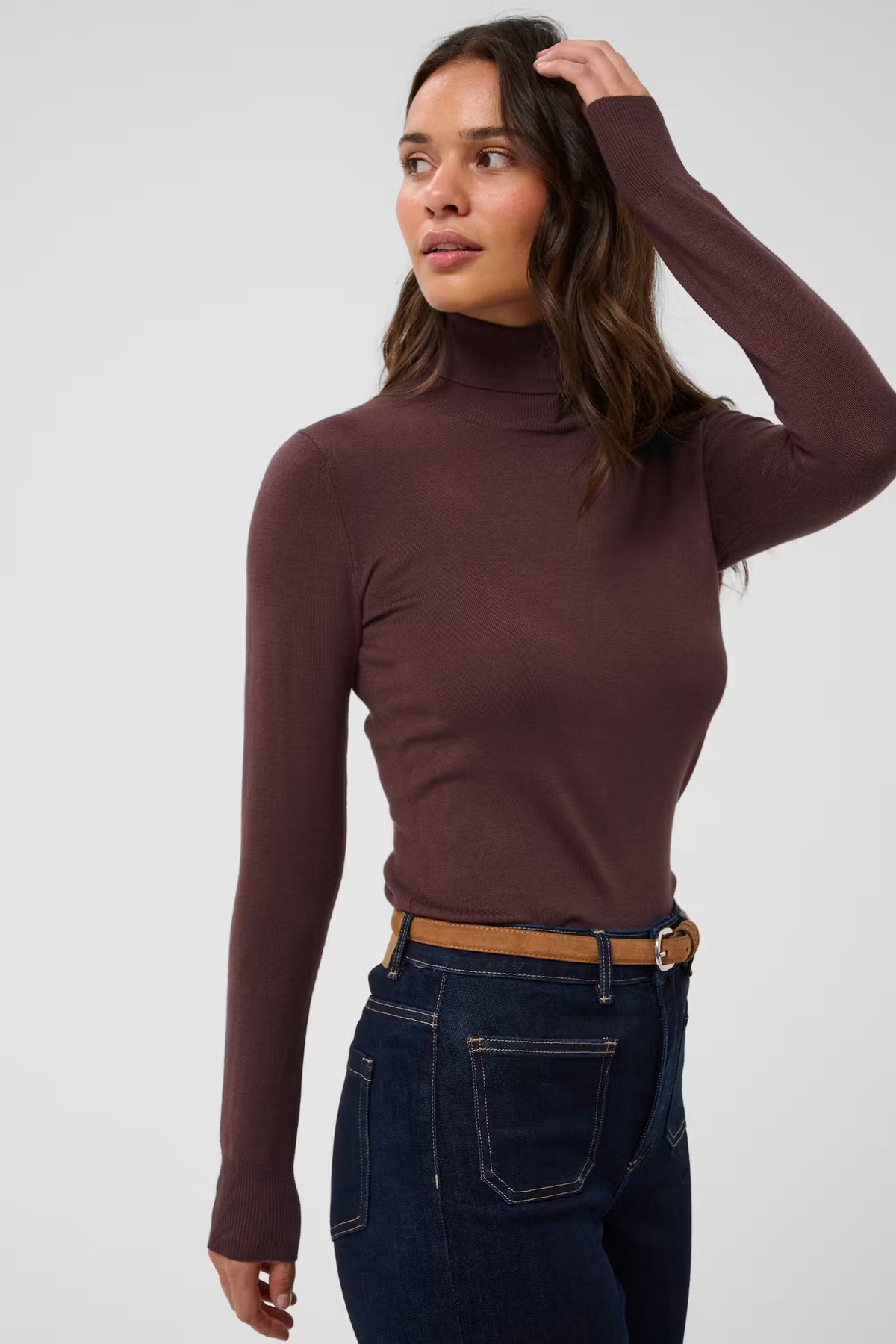 A woman wearing a fitted long-sleeve turtleneck top in a deep plum-brown color, paired with dark blue jeans and a tan belt, against a plain light gray studio background.