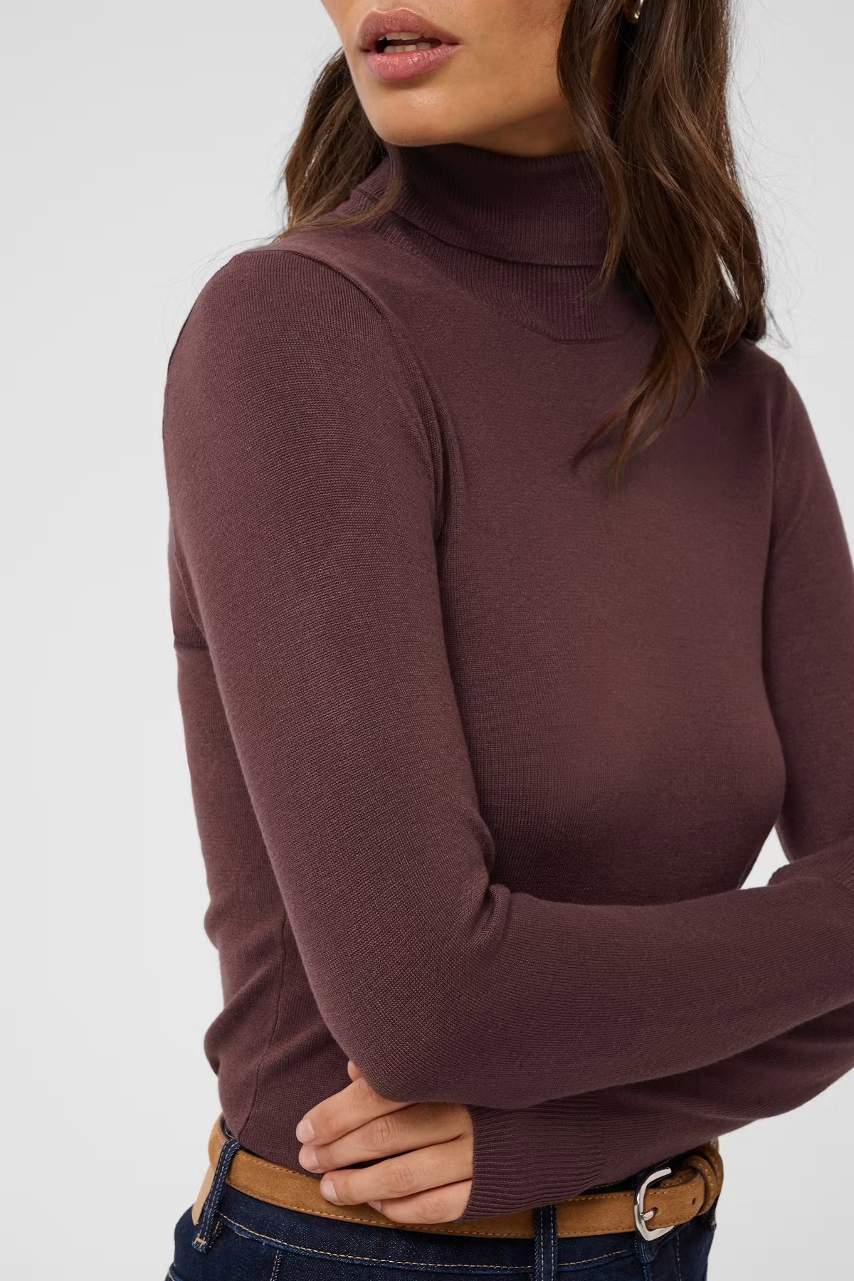 A woman wearing a fitted ribbed turtleneck sweater in a muted plum-brown color, paired with blue jeans and a brown belt, against a plain light gray studio background.