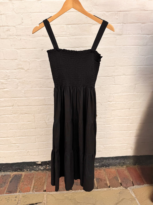 A black sleeveless midi dress with a smocked bodice and thin shoulder straps, hanging on a wooden hanger against a white painted brick wall.
