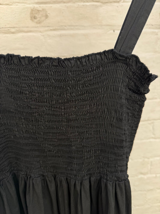 Close-up of a black smocked dress with a ruffled neckline and shoulder strap, shown against a light brick wall background.