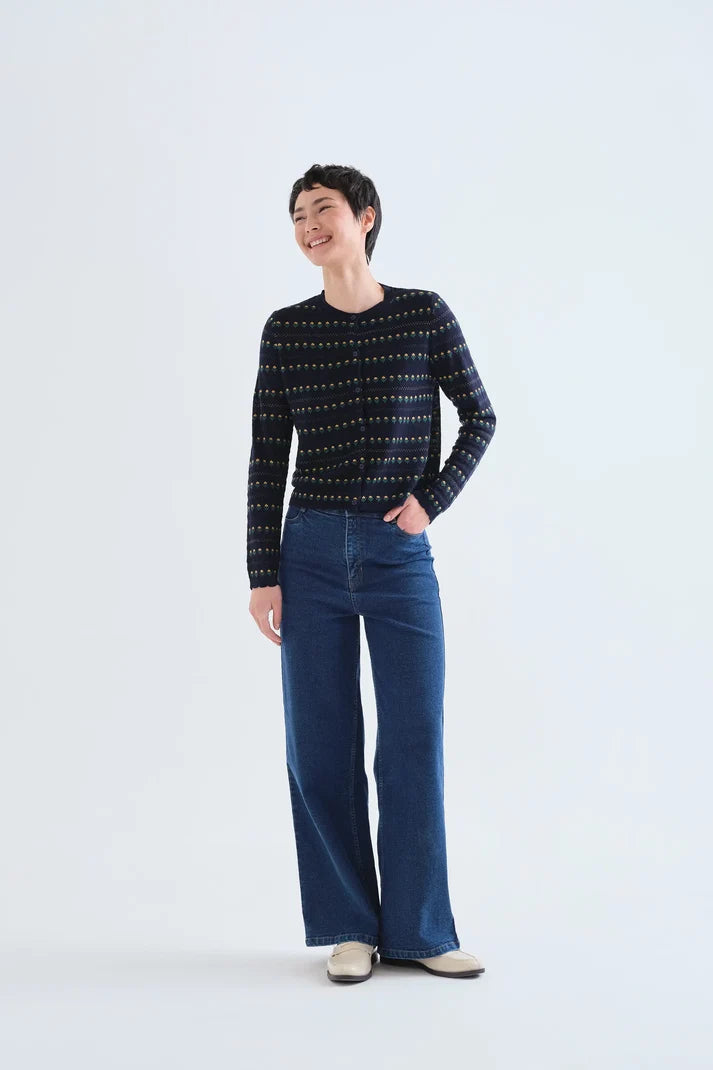 A model stands against a light gray studio background wearing a dark navy long-sleeve knit sweater with a fine geometric pattern, paired with dark blue wide-leg jeans and beige shoes.