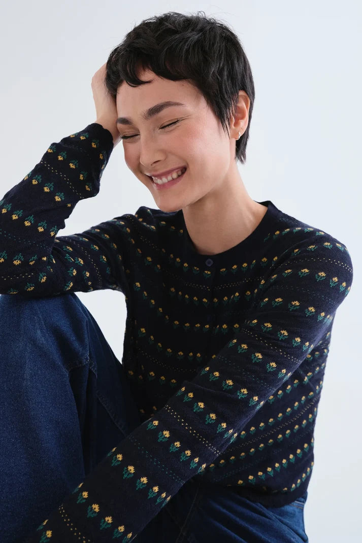 A woman with short dark hair smiling while wearing a dark navy long-sleeve knit sweater with a repeating small geometric pattern in yellow, green, and white, paired with blue jeans against a light gray background.