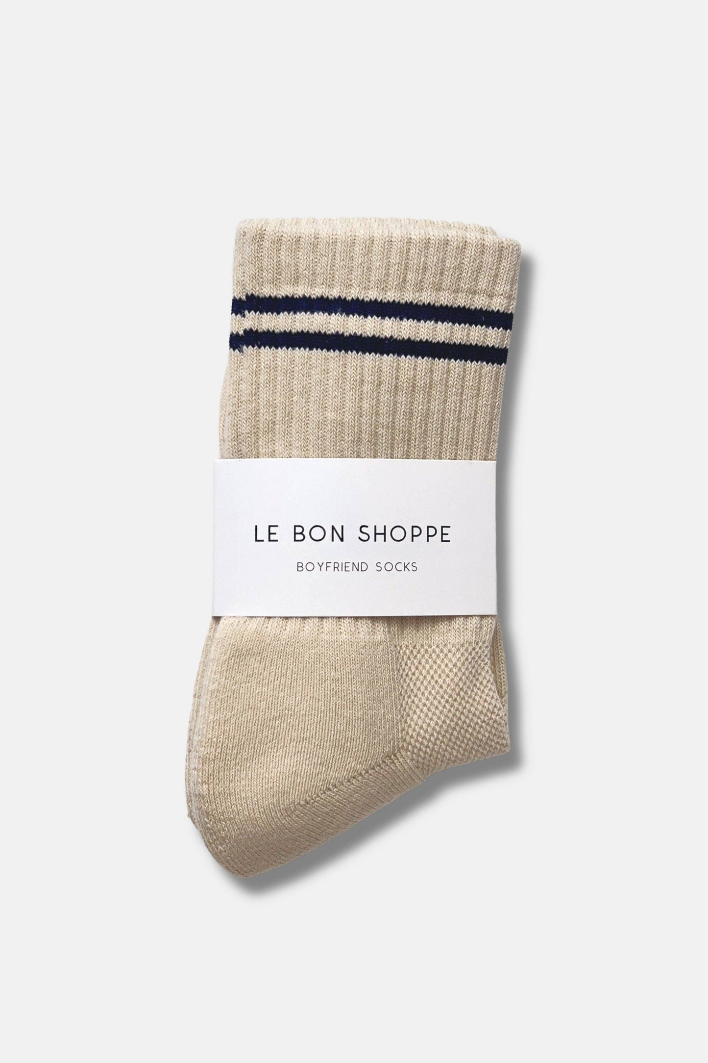 A single beige ribbed knit sock with two navy blue horizontal stripes near the cuff, wrapped in a white paper band that reads 'LE BON SHOPPE BOYFRIEND SOCKS', on a light gray background.