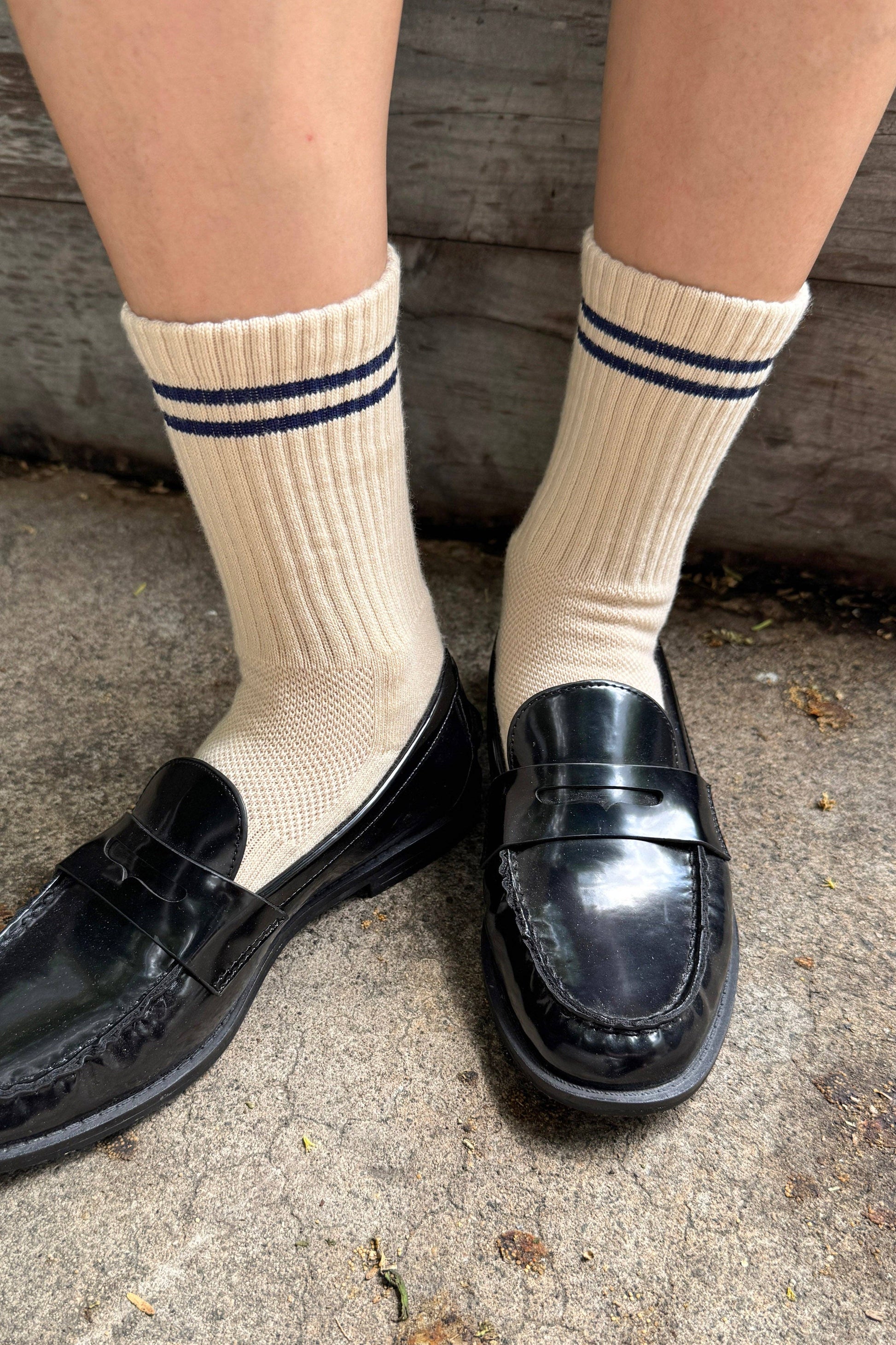 A person wearing cream ribbed crew socks with two dark navy stripes, paired with shiny black leather penny loafers, standing on a concrete surface.