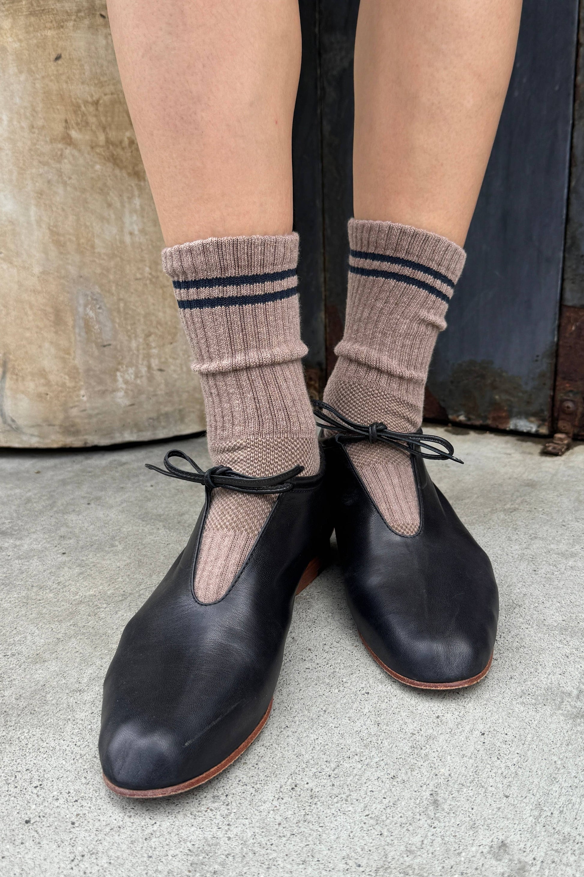 A person wearing dusty pink ribbed crew socks with navy stripes and black leather Mary Jane shoes, standing on a concrete floor.