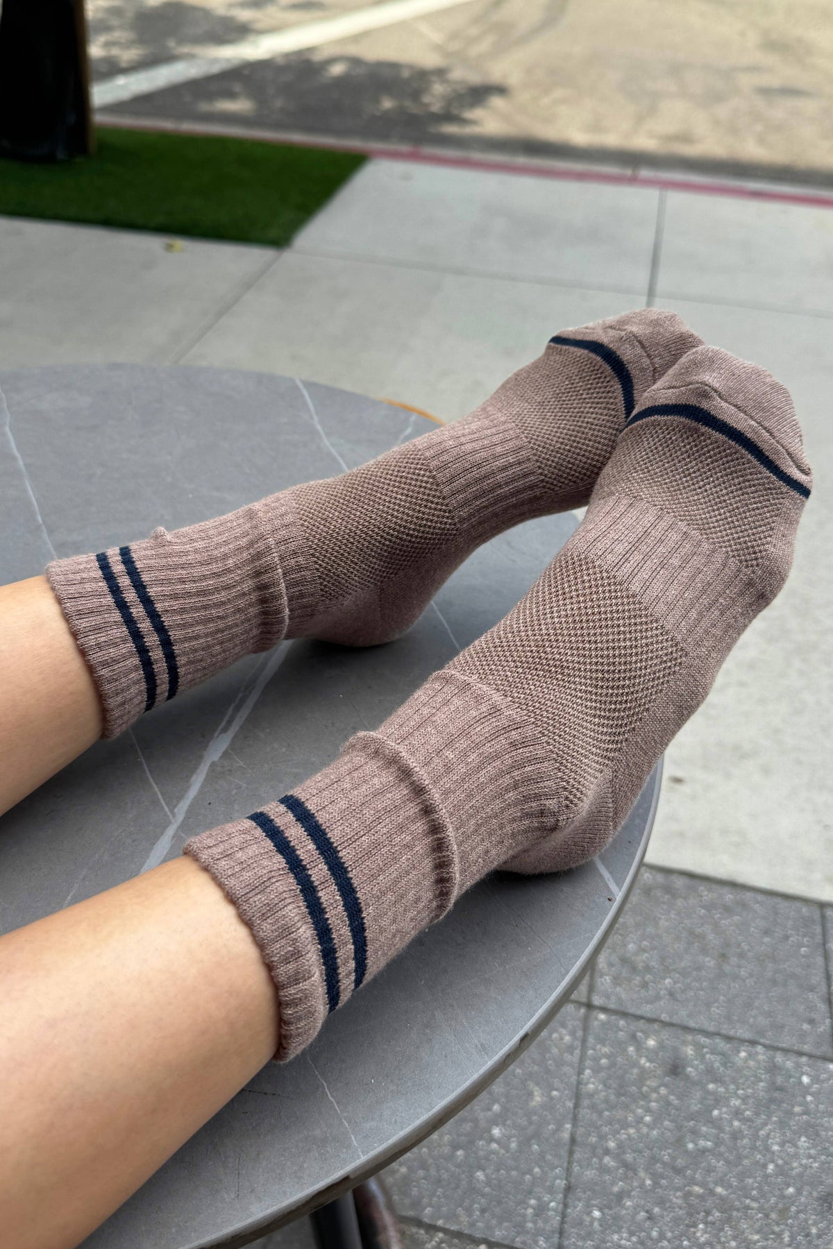 A pair of ribbed crew socks in a muted taupe-brown color with dark navy stripes near the cuffs and toes, worn on feet resting on a round gray table outdoors.