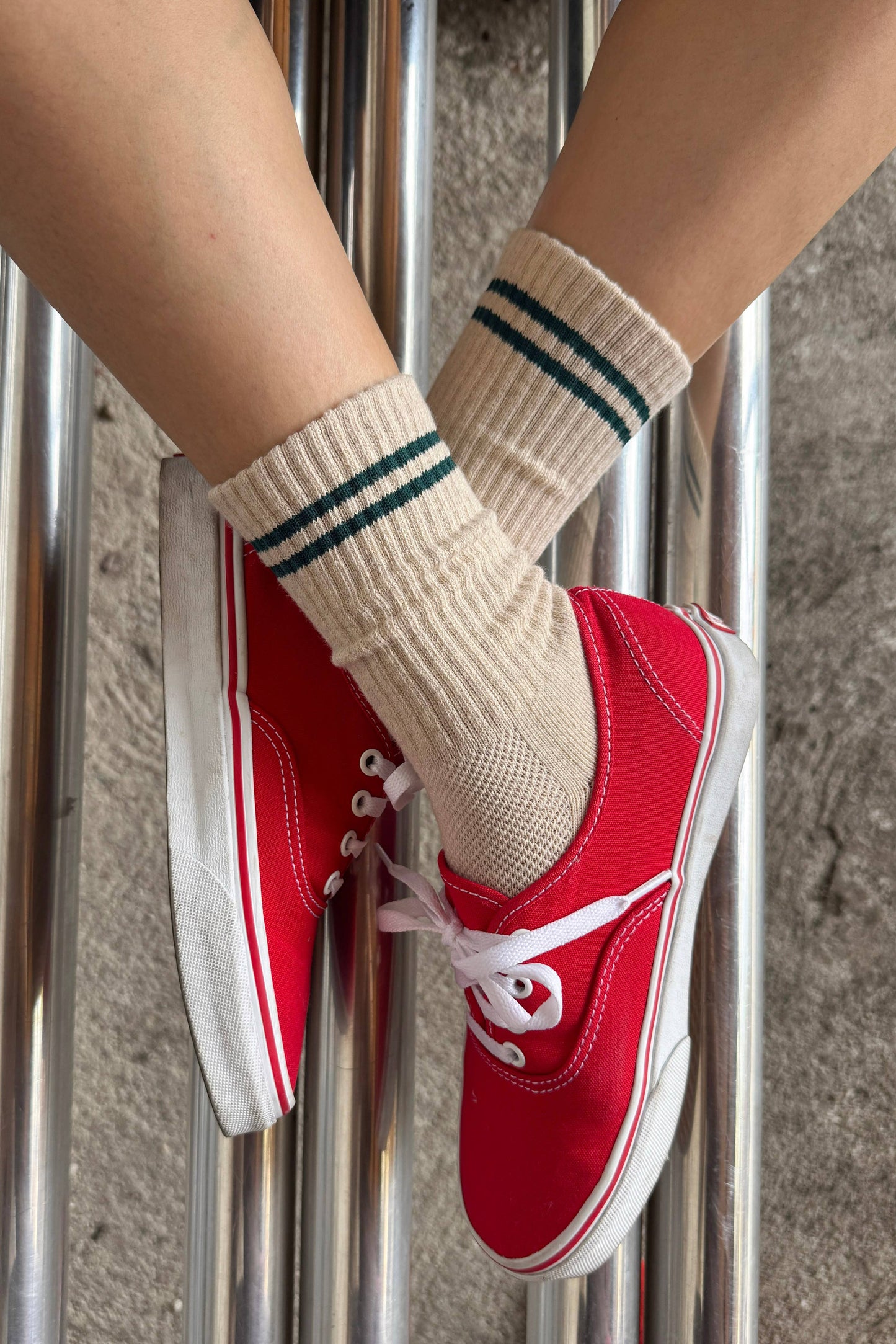 A close-up of a person's legs crossed over shiny metal poles, wearing beige ribbed crew socks with dark green stripes and red low-top canvas sneakers with white laces and soles.