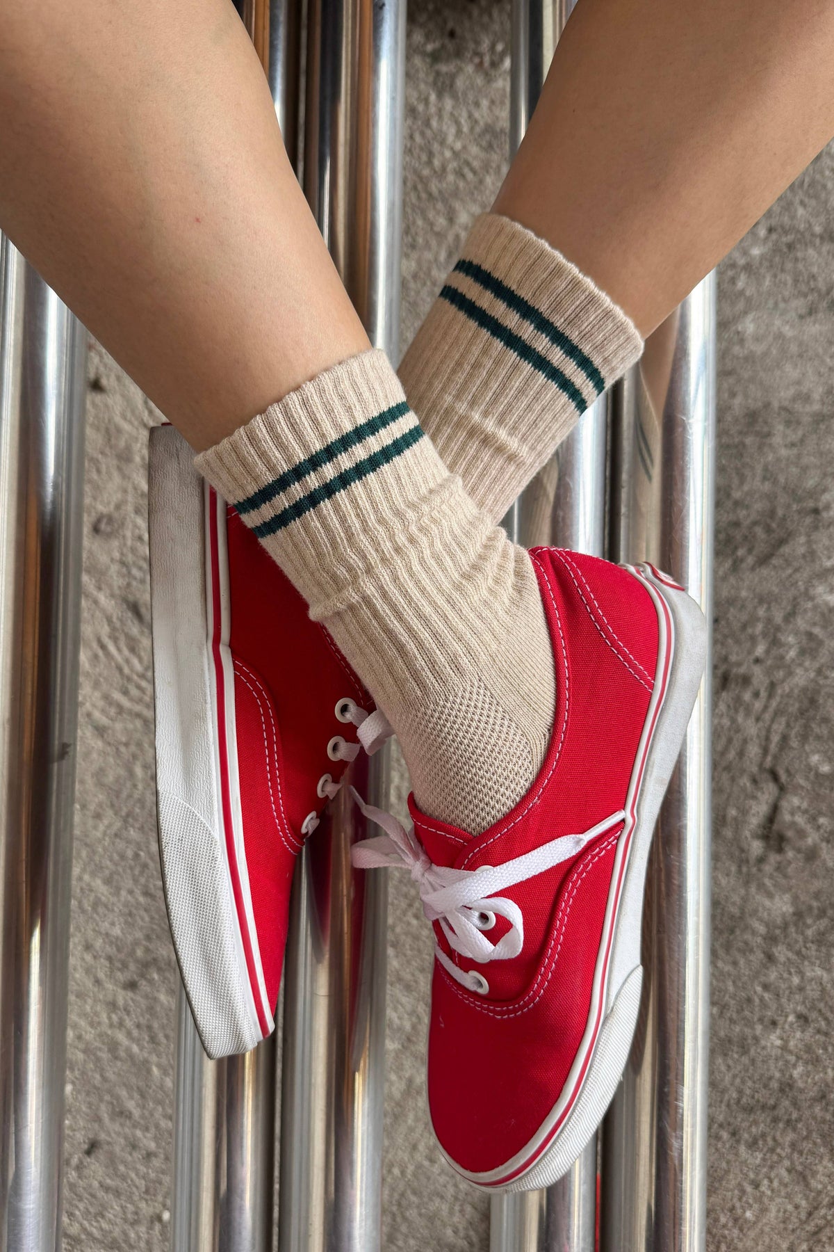 A close-up of a person's legs crossed over shiny metal poles, wearing beige ribbed crew socks with dark green stripes and red low-top canvas sneakers with white laces and soles.