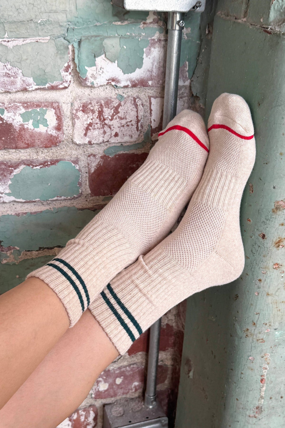 A person's feet wearing ribbed beige crew socks with dark green double stripes near the cuffs and a thin red stripe across the toes, resting against a rustic brick and painted metal wall.