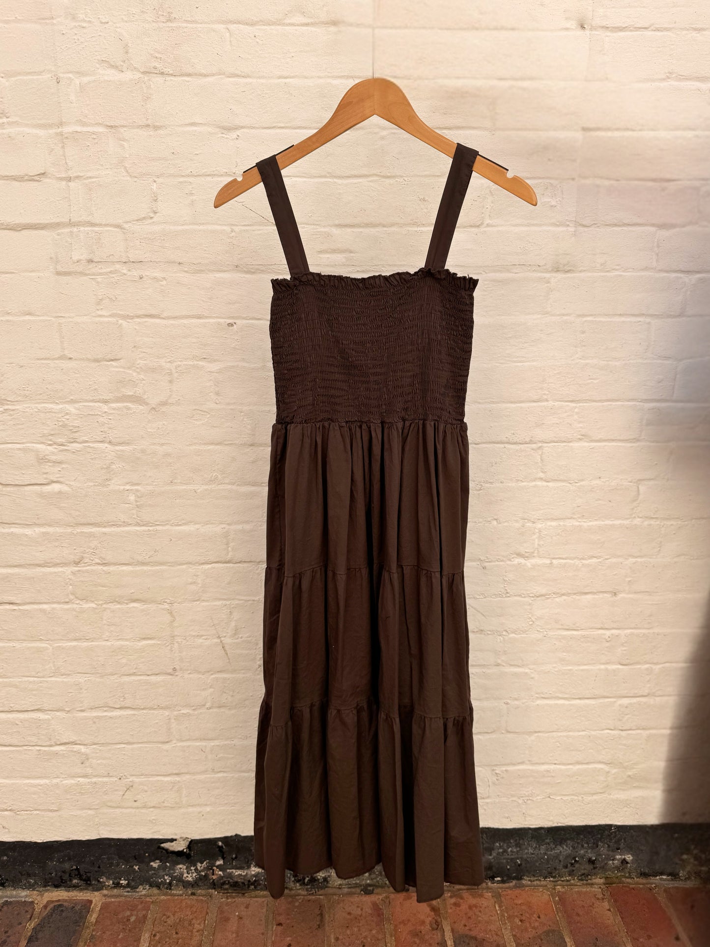 A dark brown tiered midi dress with a smocked bodice and thin shoulder straps, hanging on a wooden hanger against a white painted brick wall.