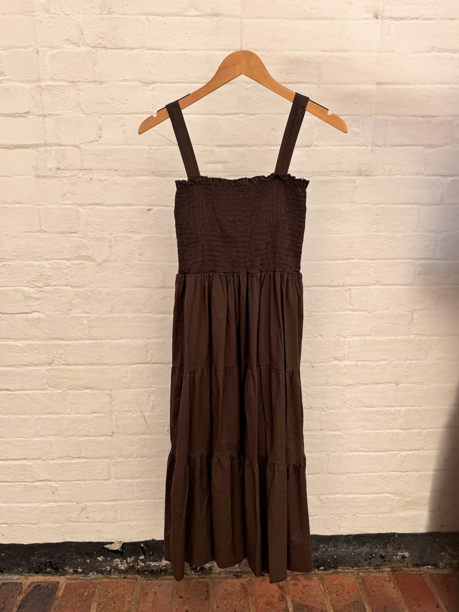 A dark brown tiered midi dress with a smocked bodice and thin shoulder straps, hanging on a wooden hanger against a white painted brick wall.