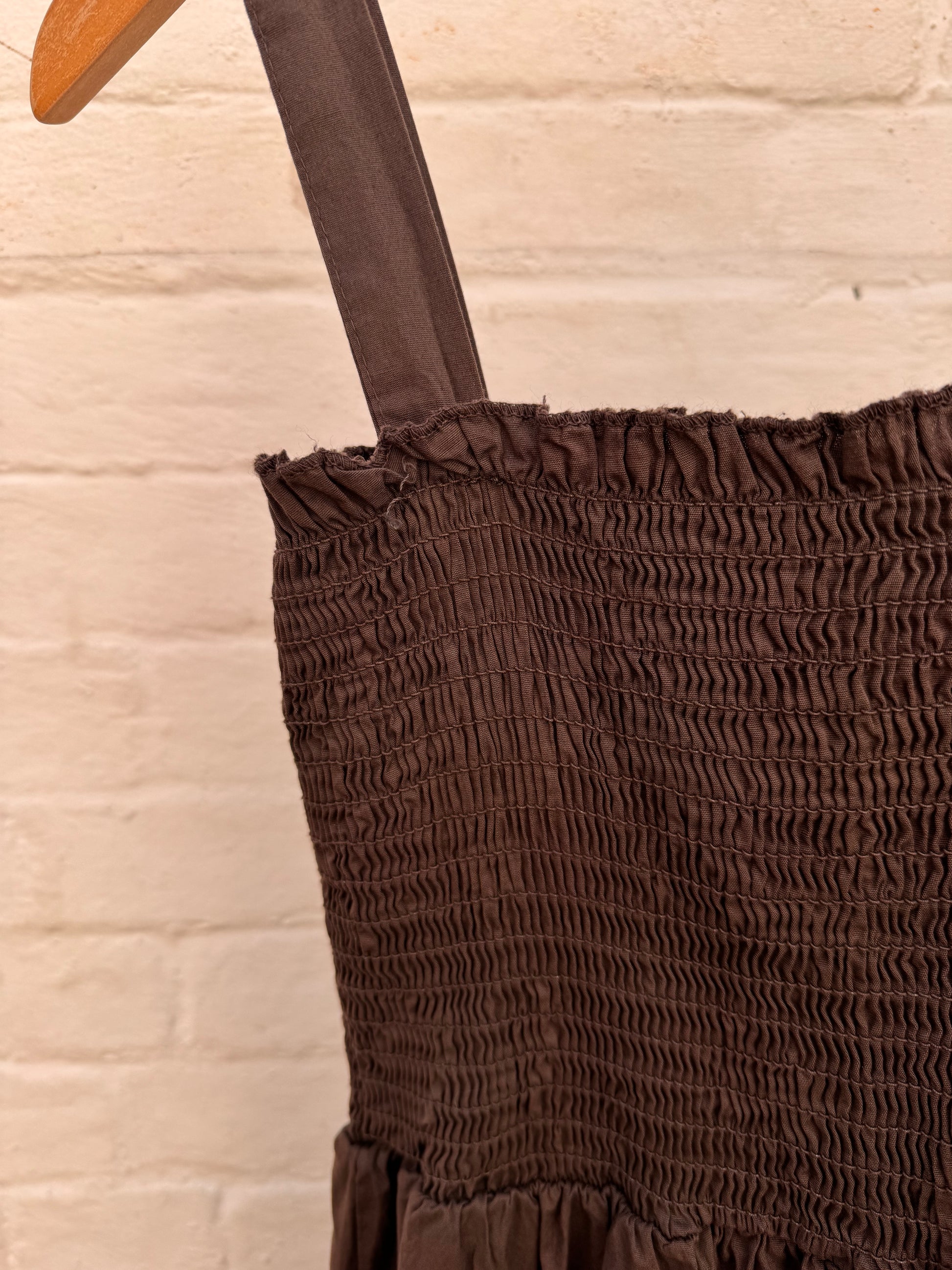 A dark brown tiered midi dress with a smocked bodice and thin shoulder straps, hanging on a wooden hanger against a white painted brick wall.