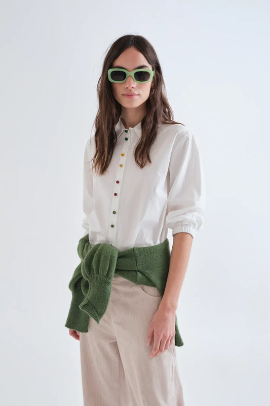 A woman modeling a white button-up shirt with colorful buttons, beige trousers, and a green knit wrap tied around her waist, set against a plain white studio background.