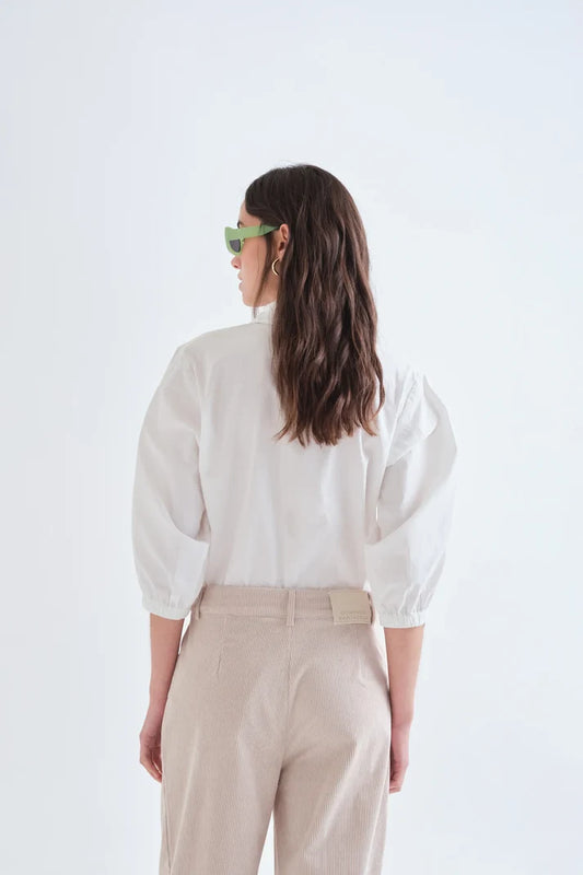 A woman is shown from behind wearing a white puff-sleeve blouse and beige trousers against a plain white studio background.