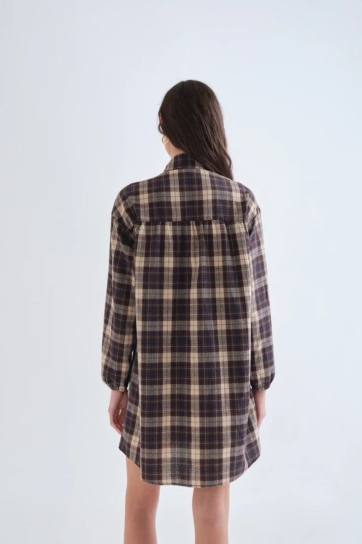 A woman viewed from behind wearing a long-sleeve plaid shirt dress in dark brown, beige, and black, set against a plain light gray studio background.