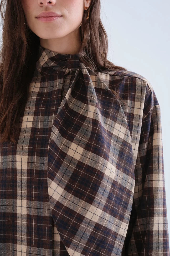 Close-up of a woman wearing a high-neck plaid blouse with a large draped tie detail at the front. The fabric features a plaid pattern in dark brown, beige, and navy blue.