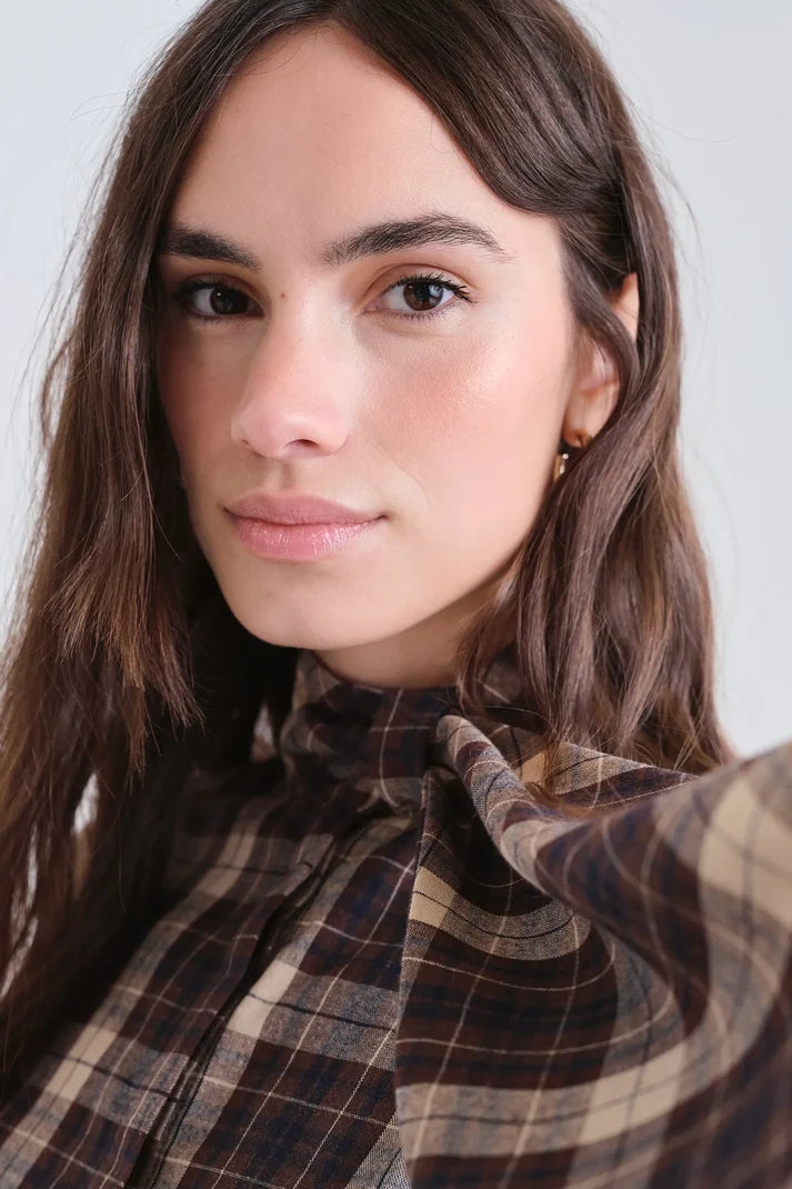 A close-up portrait of a woman with long brown hair wearing a brown and beige plaid shirt with a high neck, set against a plain light gray background.