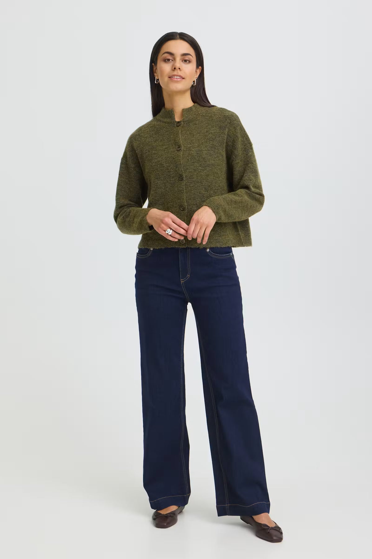 A woman modeling a cropped, olive green knit sweater with a mock neck and front zipper, paired with dark blue wide-leg jeans and dark brown flats, against a light gray studio background.