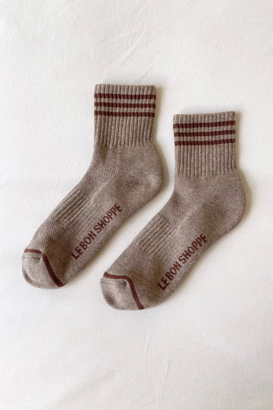 A pair of beige crew socks with dark burgundy stripes on the cuff and heel, featuring the brand name 'LERON SKOPPE' printed in burgundy on the sole, laid flat on a white textured fabric background.