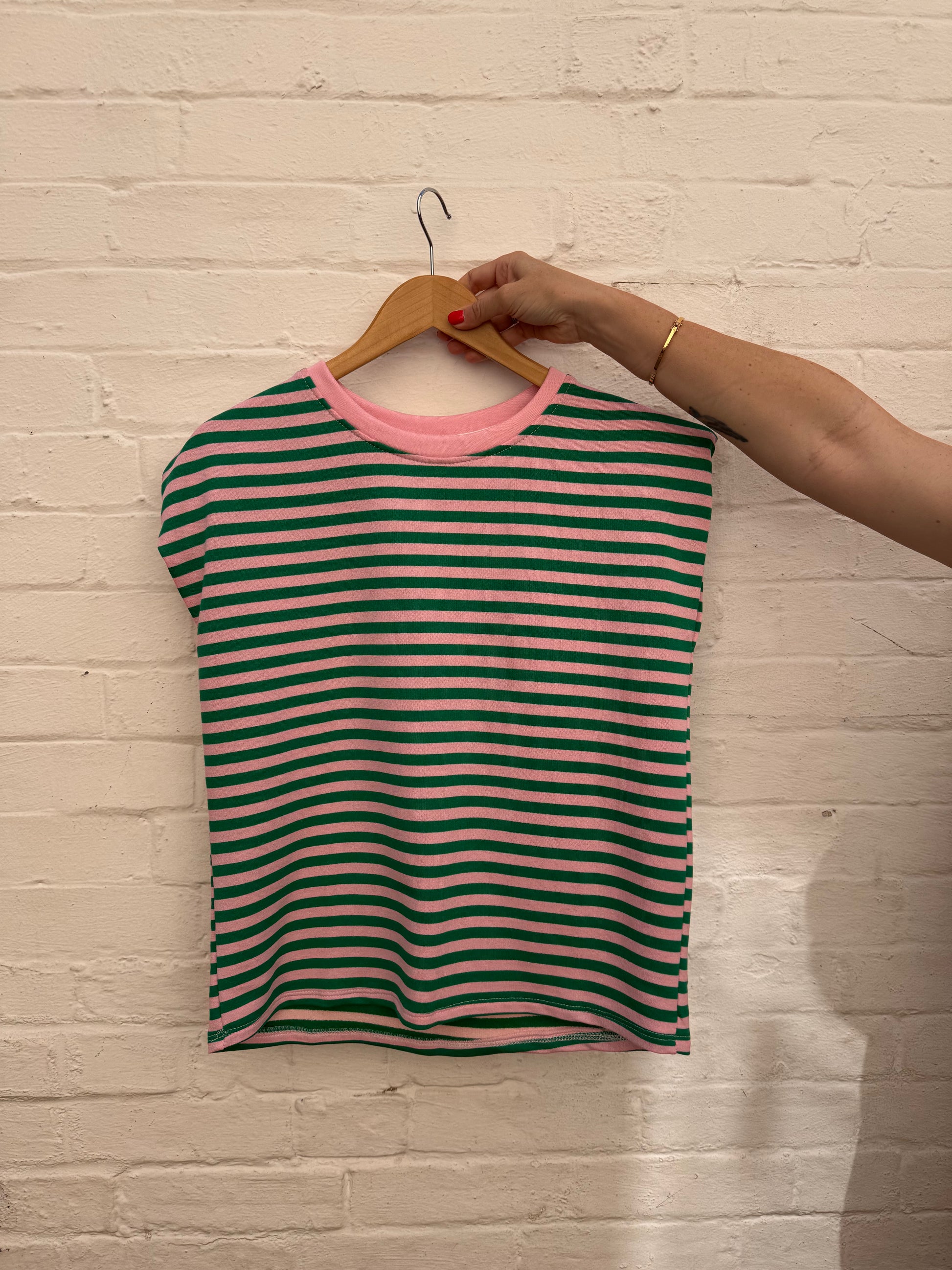 A green and pink striped short-sleeve top with a pink crew neck, hanging on a wooden hanger held by a person's hand against a white brick wall.