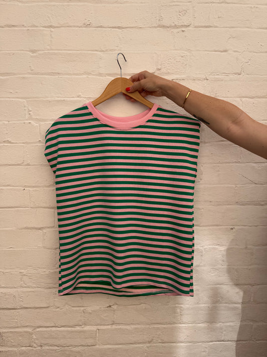 A green and pink striped short-sleeve top with a pink crew neck, hanging on a wooden hanger held by a person's hand against a white brick wall.