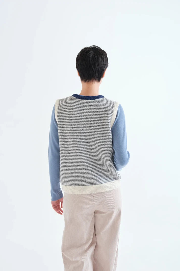 A person shown from the back wearing a gray and white striped knit sweater vest over a light blue long-sleeve shirt and beige pants, against a plain white studio background.