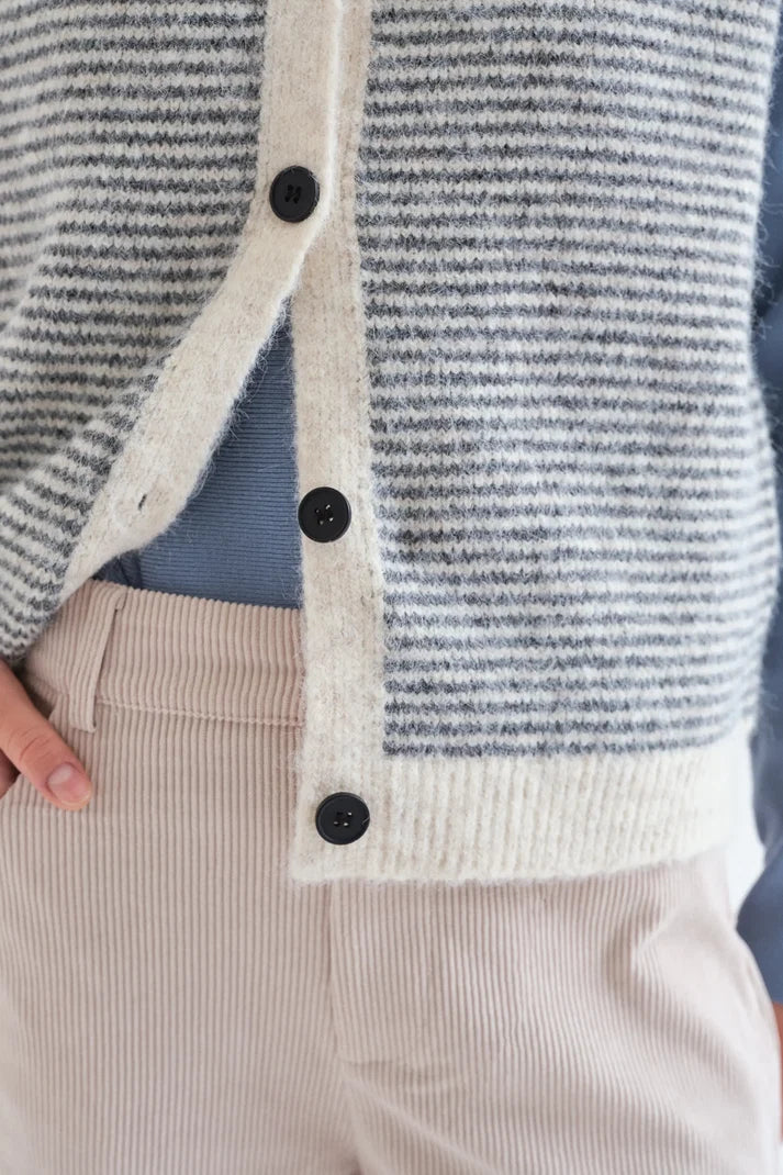 Close-up of a person wearing a fuzzy knit cardigan with gray and off-white horizontal stripes, black buttons, and a light blue top underneath, paired with light beige corduroy pants.