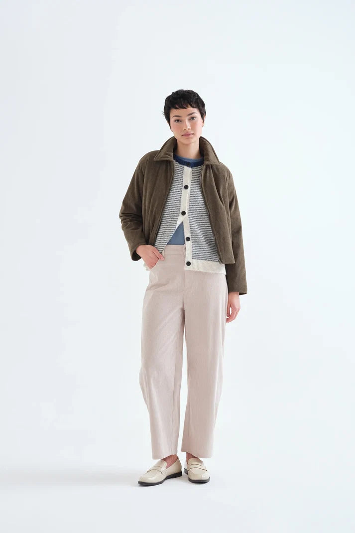 A model stands against a plain white studio background wearing a dark brown jacket over a gray and white striped button-up shirt, paired with light beige wide-leg trousers and cream-colored shoes.