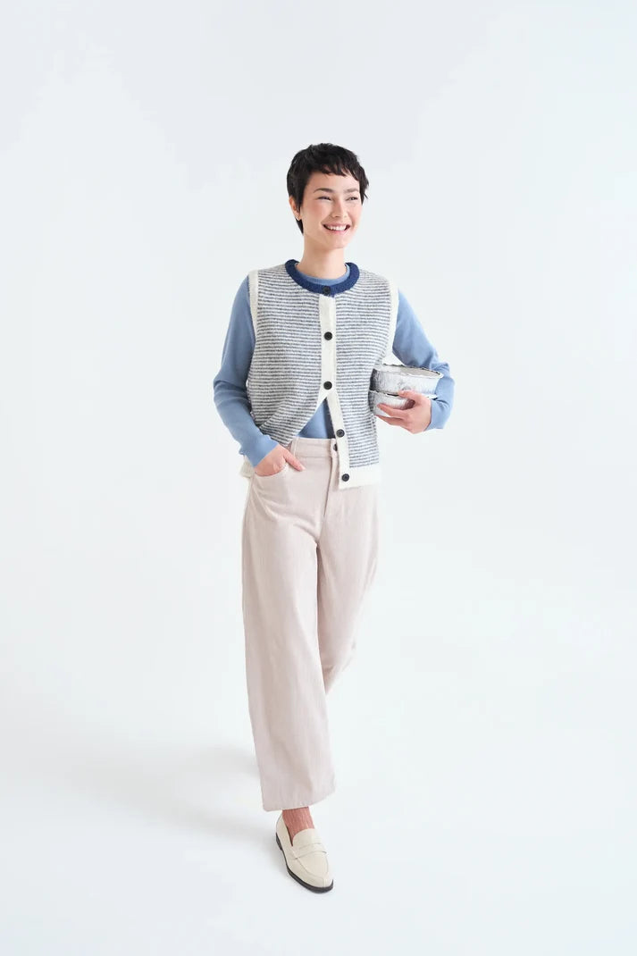 A woman modeling a light gray and white textured knit cardigan vest over a blue long-sleeve shirt, paired with beige wide-leg trousers and cream loafers, holding a small silver bowl against a plain white studio background.