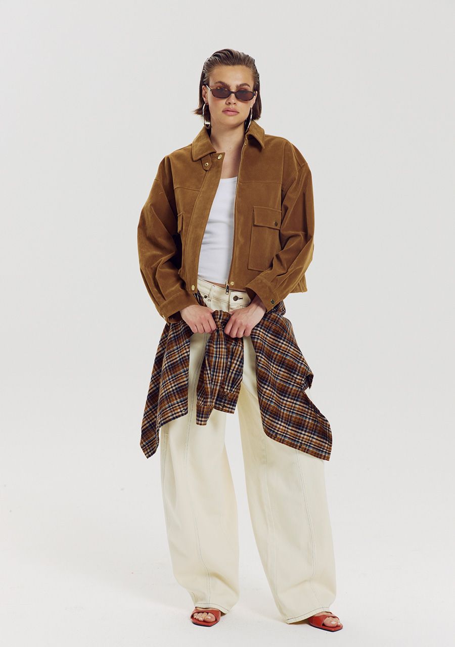 A model stands against a light gray studio background wearing a brown corduroy jacket over a white top, cream wide-leg pants, sunglasses, and red sandals. A plaid shirt is tied around the waist.